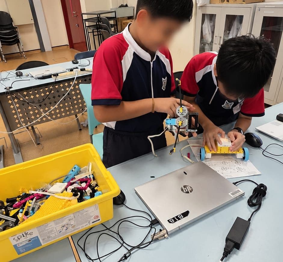 Two students building a SPIKE Prime model, with the parts bin, a build sheet and a school laptop on the table