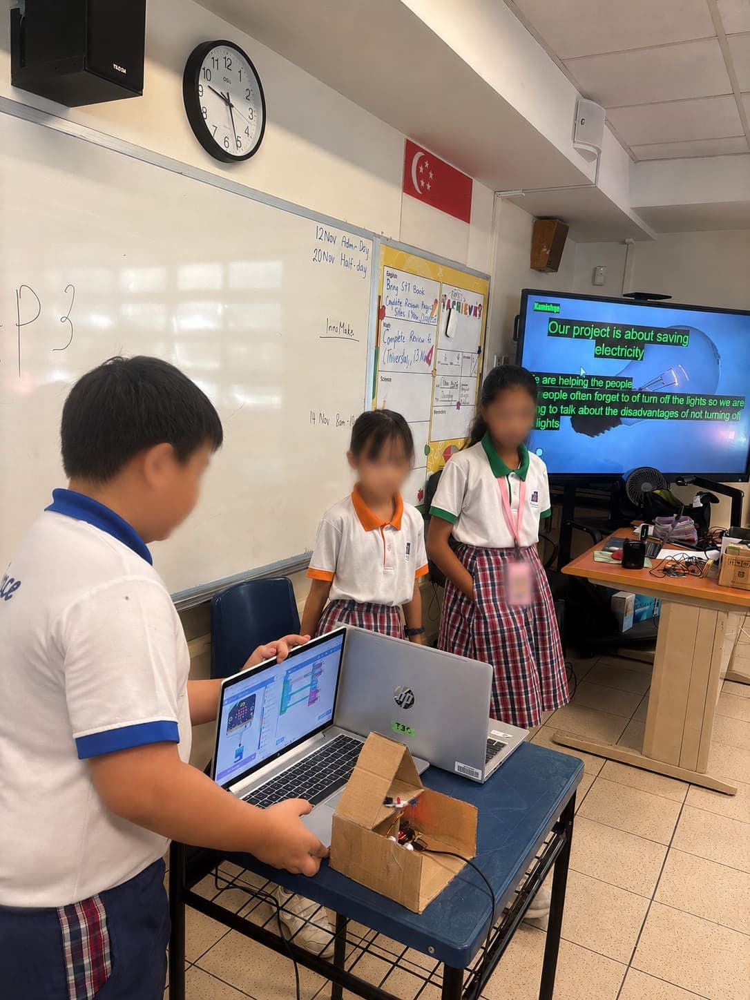 Primary students presenting a micro:bit project about saving electricity, with their cardboard build on the desk