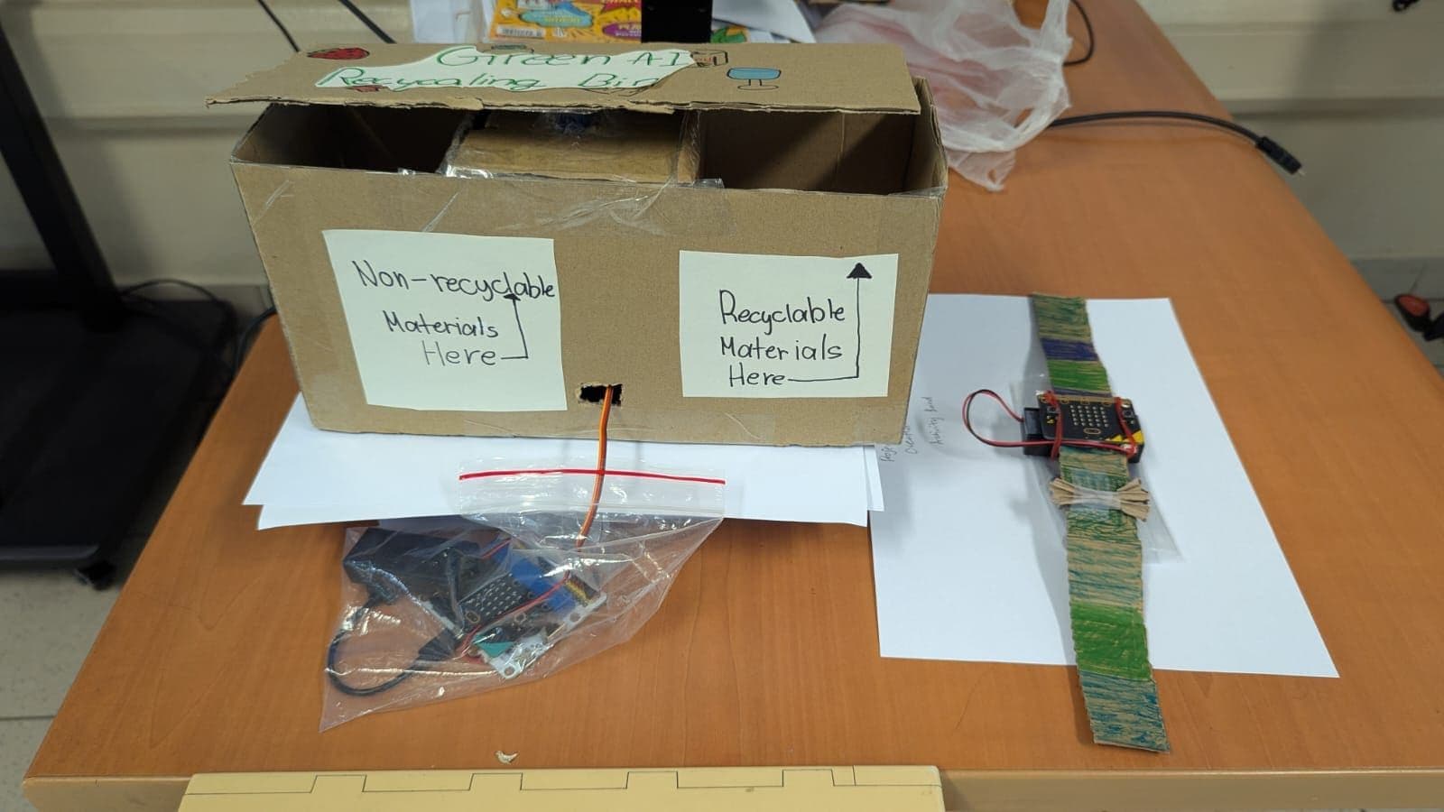 A cardboard recycling bin prototype wired to a micro:bit, beside a micro:bit wearable band