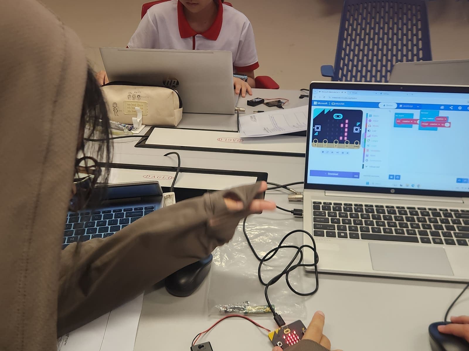 MakeCode block editor open on a laptop beside a micro:bit lighting up in a student’s hand